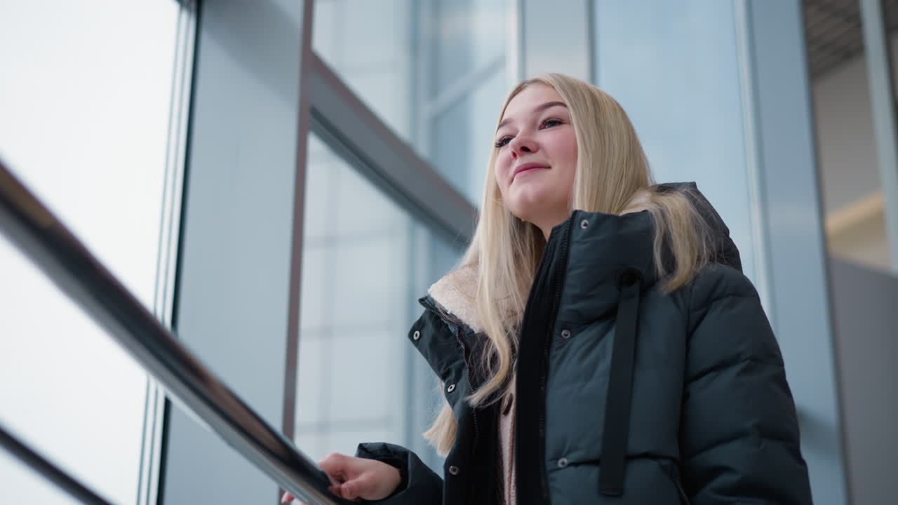 Donna in giacca con uno sguardo contemplativo e dolce vicino a un edificio di vetro, che emana una tranquilla sicurezza, i suoi lunghi capelli biondi le ricadono sulle spalle mentre appare immersa nei suoi pensieri.