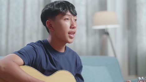primer plano de un compositor asiático con un cuaderno en la mesa cantando y tocando la guitarra en casa