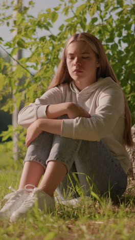la mujer se sienta en el suelo césped con los ojos cerrados, descansando contra el tronco del árbol, la luz del sol se refleja en su rostro pacífico, sus brazos están plegados sobre sus rodillas, con los postes de fondo borrosos y el estadio