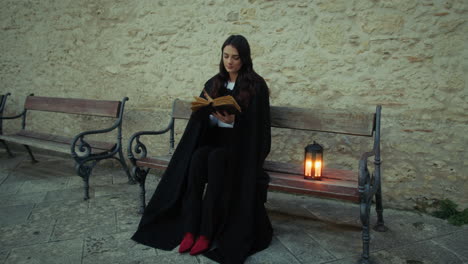 woman with cloak reads the mysteries in the book sitting on a bench in the city