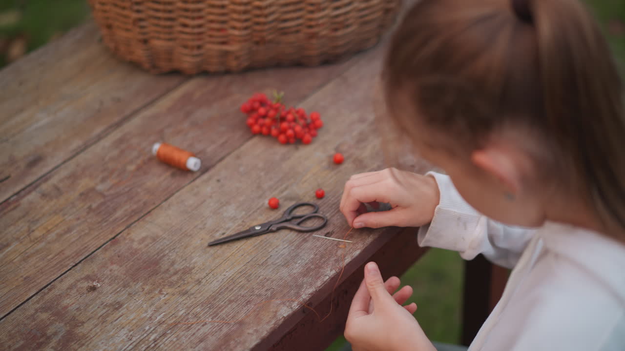 Young white girl picking red berries on wooden table with basket and scissors, hands threading berries near spool of thread, rustic outdoor garden setting, calm nostalgic mood, child maker creating