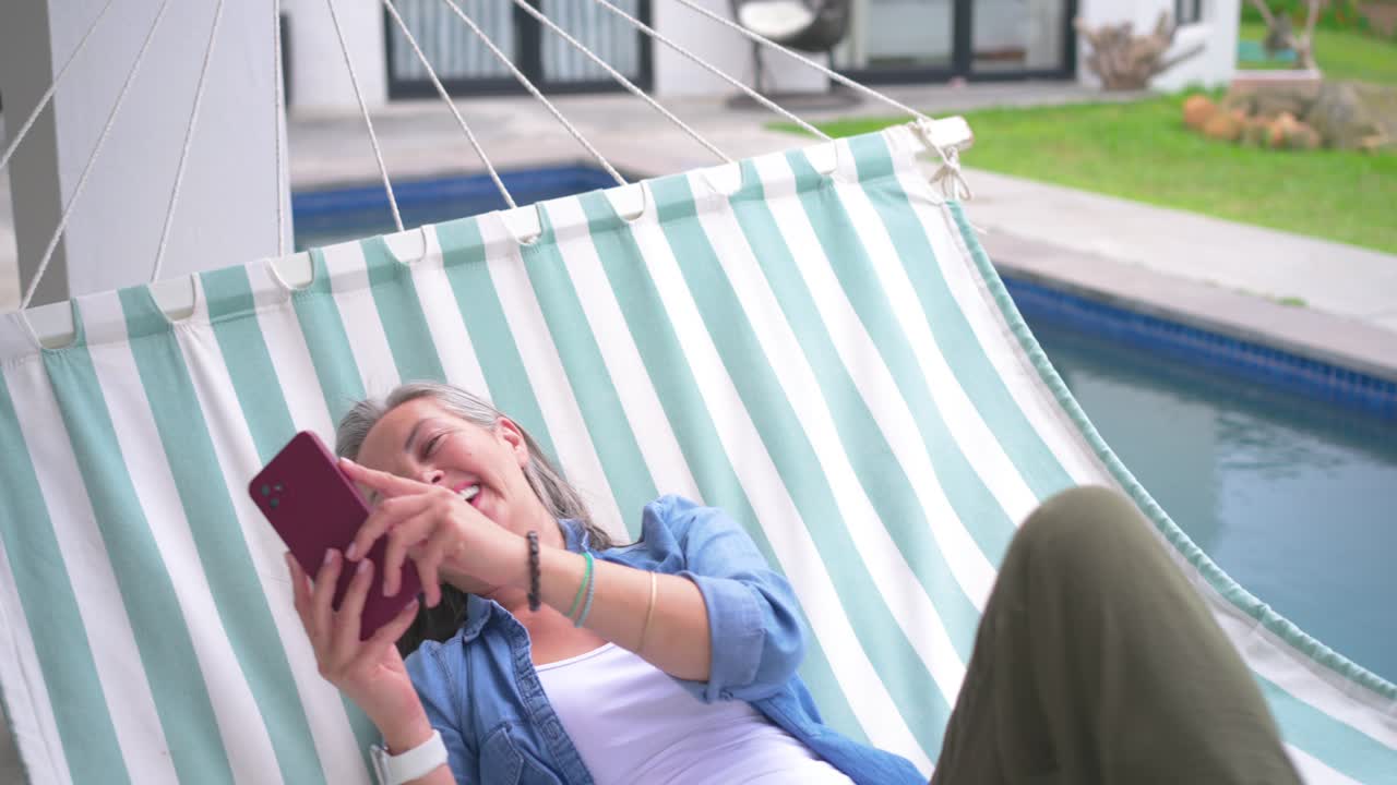 Téléphone s'illuminant avec un message entrant, femme âgée faisant défiler l'écran et riant dans un hamac au bord de la piscine.