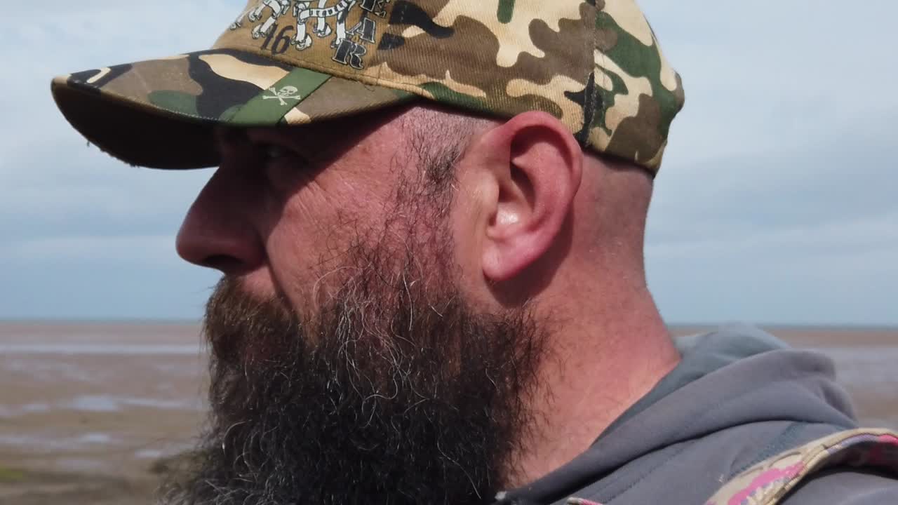 hombre urbano barbudo que se siente estresado usando una gorra de béisbol caminando por la playa ventosa