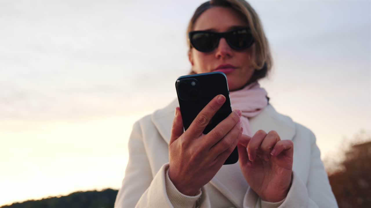 Donna elegante con cappotto bianco, sciarpa e occhiali da sole che controlla il telefono mentre è in piedi su una spiaggia al tramonto