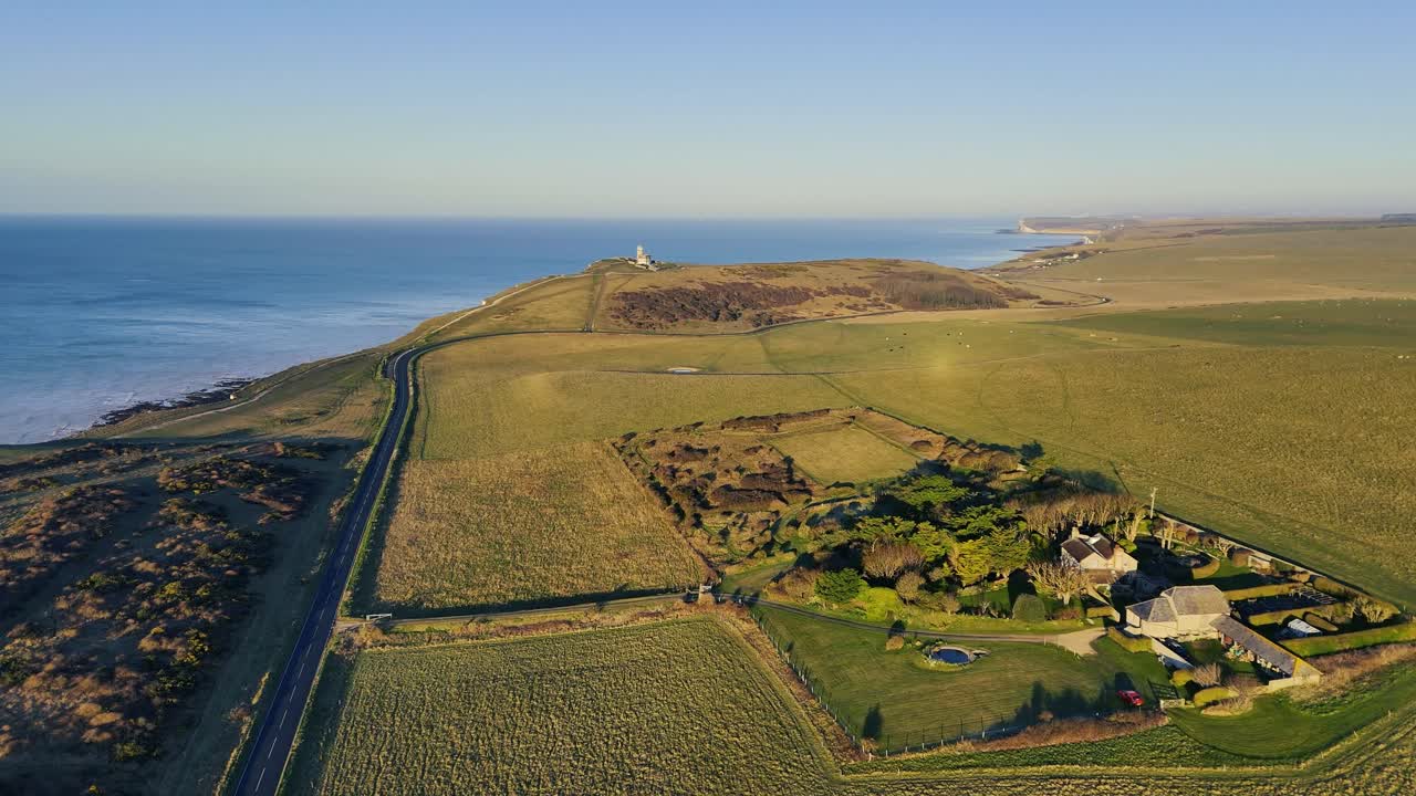 Aerial footage of a lighthouse approach, flying over a cliff-top road, farm, and lush green fields. Stunning coastal views perfect for travel, nature, and landscape projects.