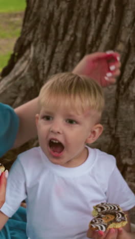 la familia sentada bajo un gran árbol, el niño más pequeño abre emocionado la boca para tomar un bocado jugoso de su bocadillo, mientras la madre mira, sosteniendo otro bocadillo