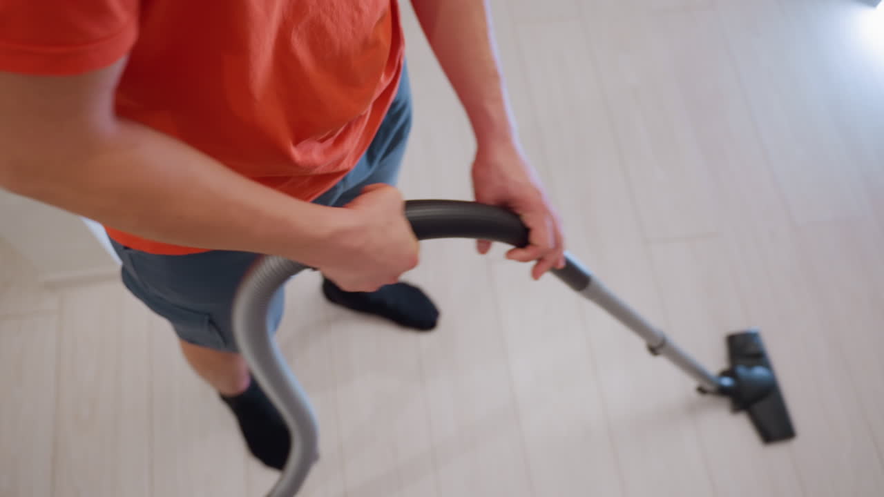Top down view of man in orange shirt and shorts holding vacuum cleaner with two hands while cleaning light wooden floor, showing daily household task, hygiene maintenance