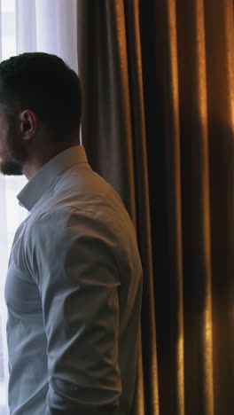 Handsome man after the work in apartment. Young businessman near the window in dark hotel room. Man arrived to a hotel. Vertical video