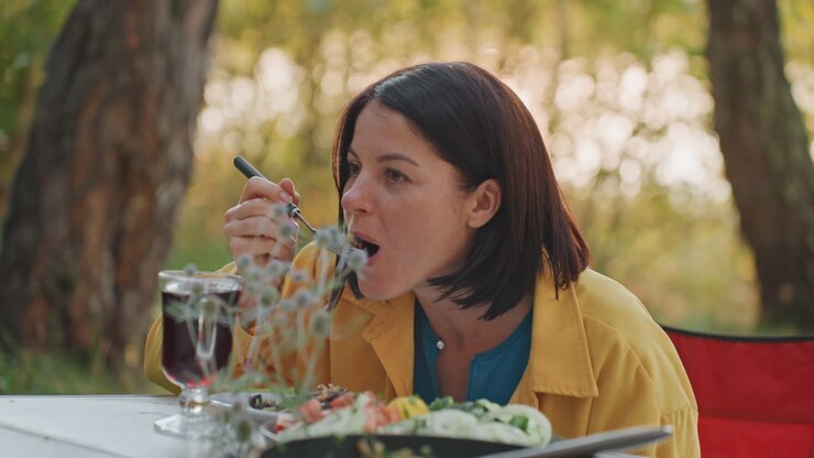 White woman camping dinner at table, lakeside picnic setting with yellow jacket, wine glass, skillet, plate and fork, travel writer savoring homemade meal, warm sunset light, calm forest background