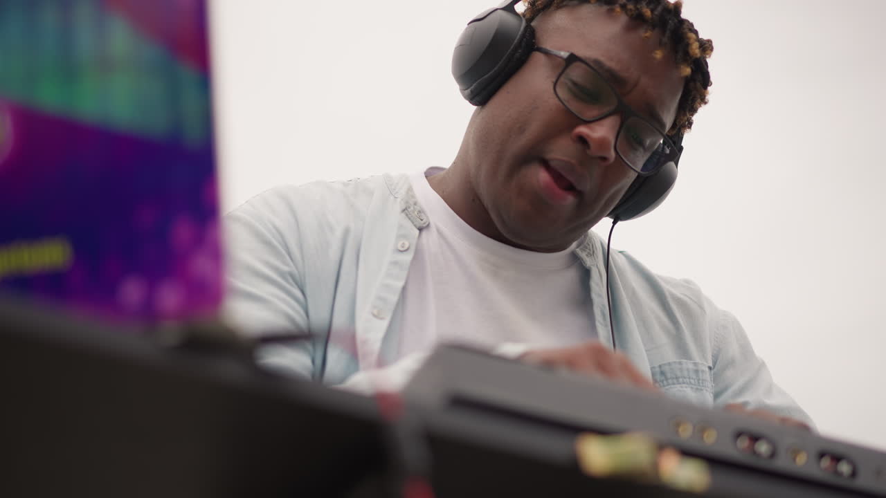 Black DJ performing energetic outdoor set with headphones and controller, dynamic gestures over mixer, engaged crowd energy, festival signage in background, headliner vibe and lively soundmix