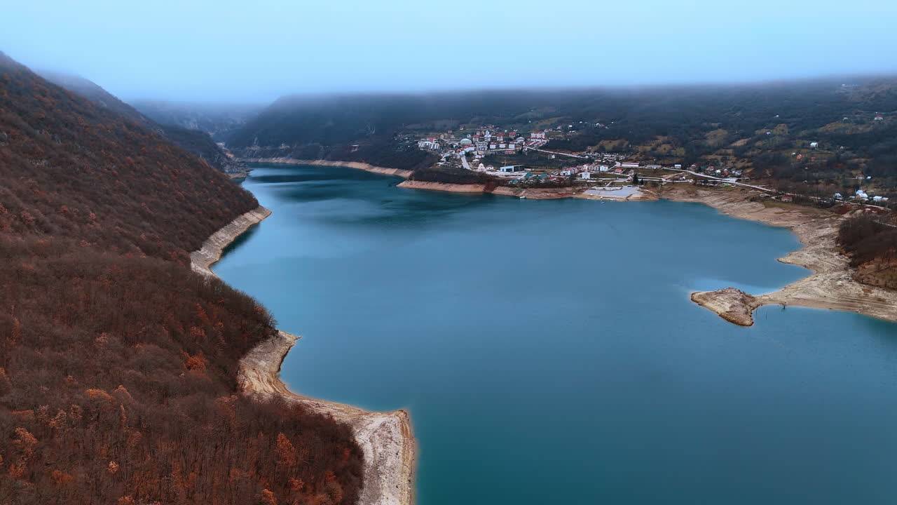 Luchtfoto van het Pivameer en een bergdorp in Montenegro. Bovenaanzicht van een turkoois stuwmeer omgeven door bergen en een kleine nederzetting onder een mistige hemel.
