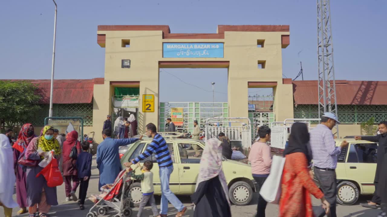 Lively atmosphere of a crowded market entrance in Islamabad, Pakistan, teeming with people from all walks of life browsing stalls, shops, and street vendors.