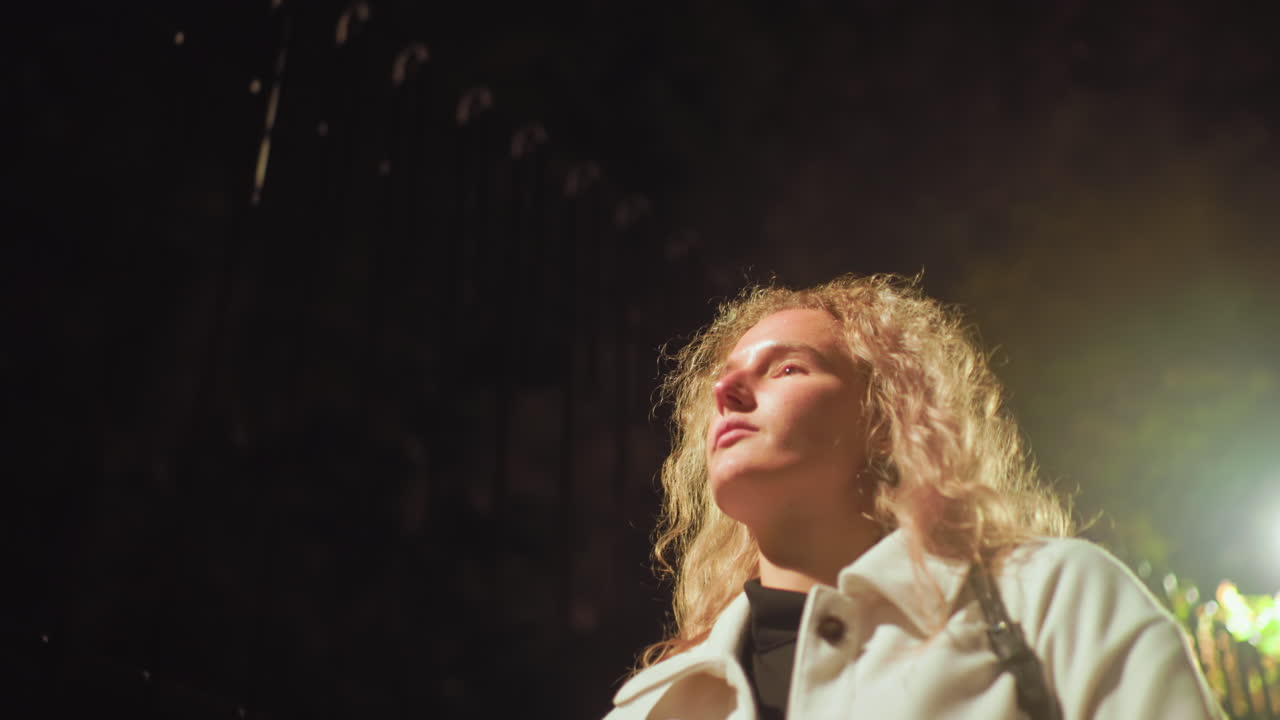 Woman with curly hair in white coat walking at night, eyes closed, calm expression on face, illuminated by bright streetlight creating dramatic glow and soft shadows against dark urban background