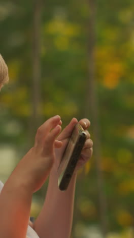 blond boy enjoys pastime with smartphone on balcony. adorable child focuses on interesting game sitting in armchair with breathtaking view of countryside