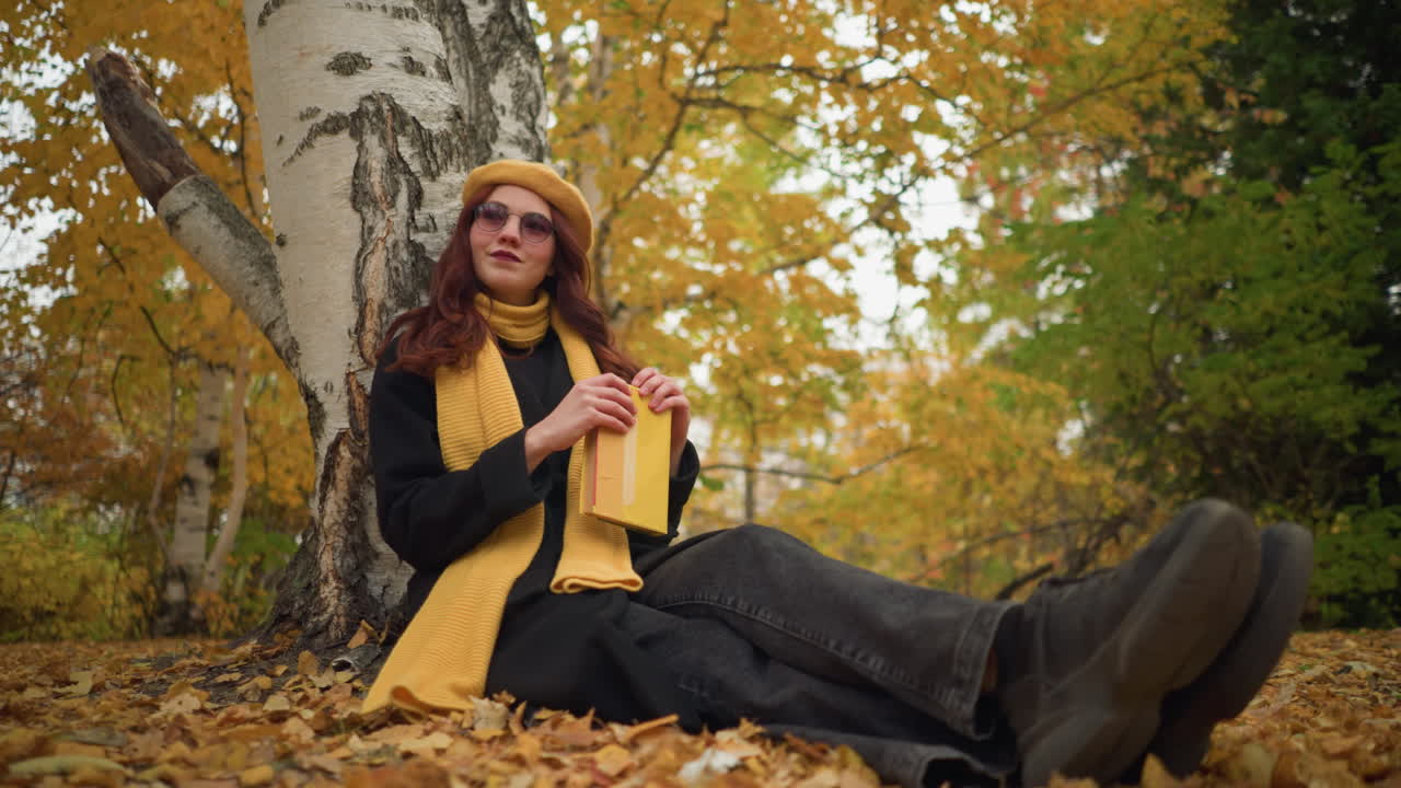 прекрасная дама в желтом берете сидит на земле, закрывает книгу, опирается на нее рукой с теплой улыбкой в спокойном парке, окруженном осенней лиственницей, наслаждаясь спокойным моментом в природе, излучающей удовлетворение.