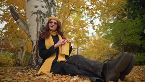 fair lady in yellow beret sits on ground, closes book, rests hand on it with warm smile in serene park, surrounded by autumn foliage, enjoying peaceful moment in nature radiating contentment