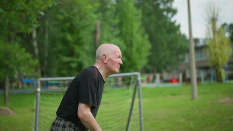 an elderly man skillfully heads a soccer ball away, focusing intently with a blurred view of a goalpost, trees, and a building in the background