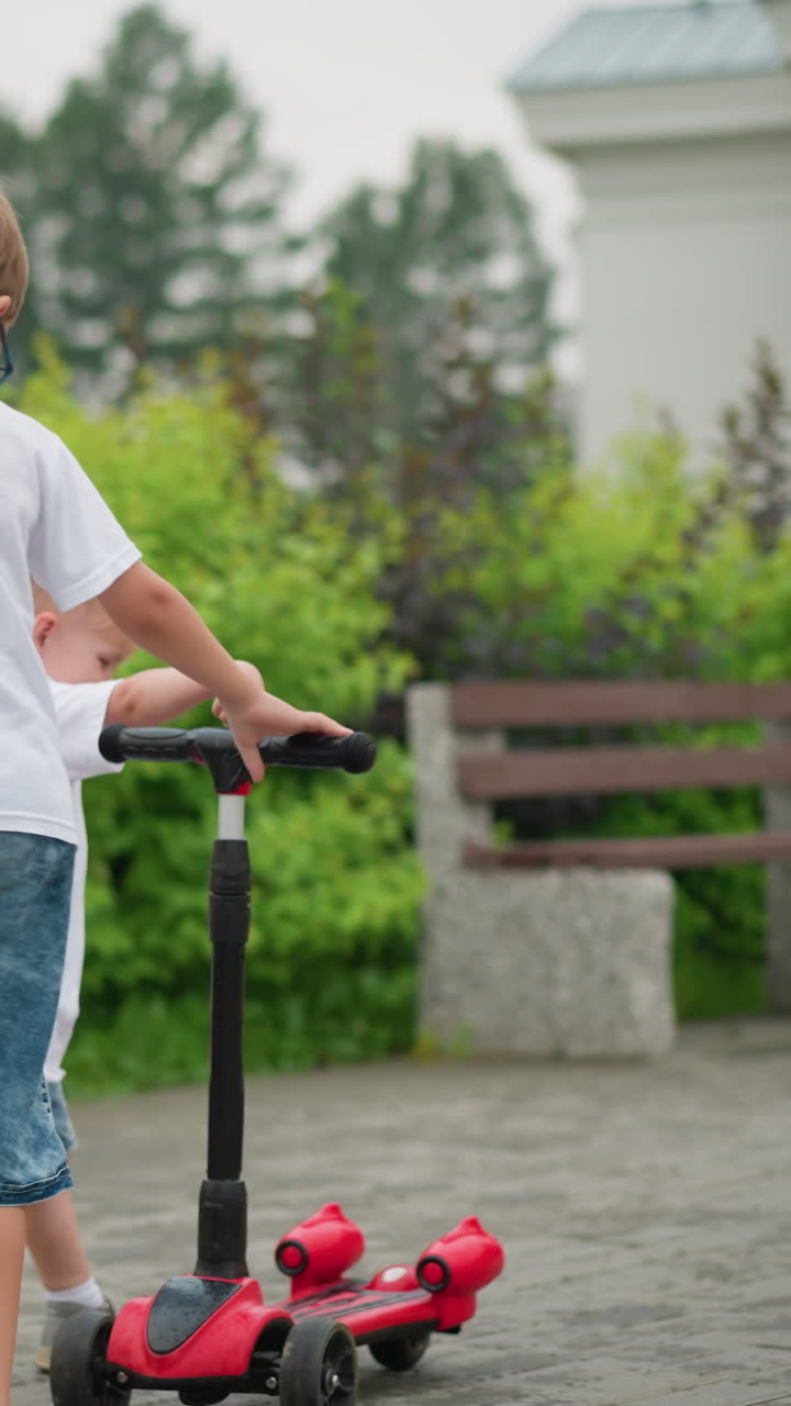 A younger and older brother joyfully run around a scooter outdoors, both holding onto the handlebar as they play together