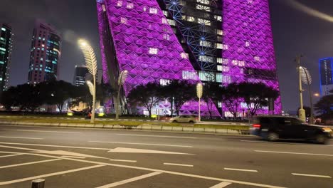 night view of a purple-lit skyscraper and city street with traffic