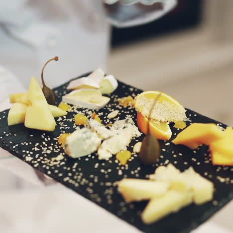 a waiter is holding in his hand a dark wooden board with different types of hard cheese decorated with fruit and sprinkled with sesame in the restaurant. cheese tasting.