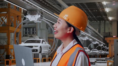 vista lateral de cerca de una ingeniera asiática con casco de seguridad trabajando en una computadora portátil y mirando a su alrededor mientras está de pie en una fábrica de automóviles ev