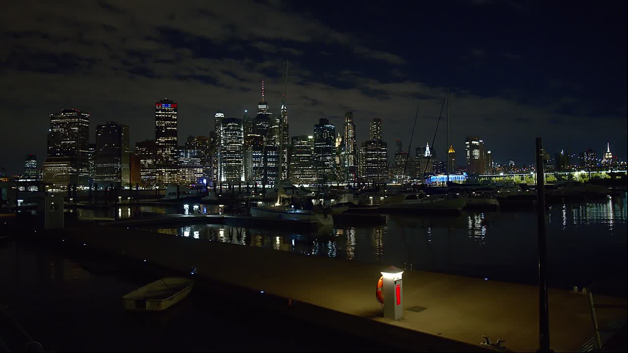 Wide shot of Brooklyn Bridge docks at night with NYC skyline in background and moored boats