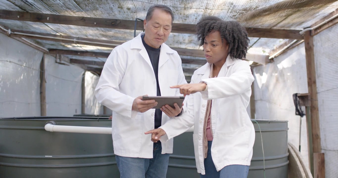 Senior Asian man and African woman in lab coats pointing at tanks tapping tablet correlating piping