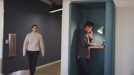 millennial woman walks past a man using a laptop and phone in a private booth in an office space