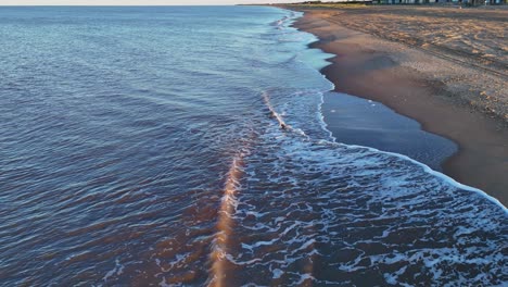 Gentle-incoming-North-Sea-tide-gently-caresses-the-golden-sandy-shoreline-with-foaming-waves-lapping-the-beach-as-the-brightly-lit-sun-begins-to-set-in-the-distance