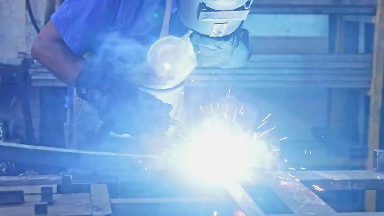 ootage of a welder in an aged workshop, wearing protective gear, as he welds metal, with tools, equipment, and ambient light creating a rugged industrial atmosphere