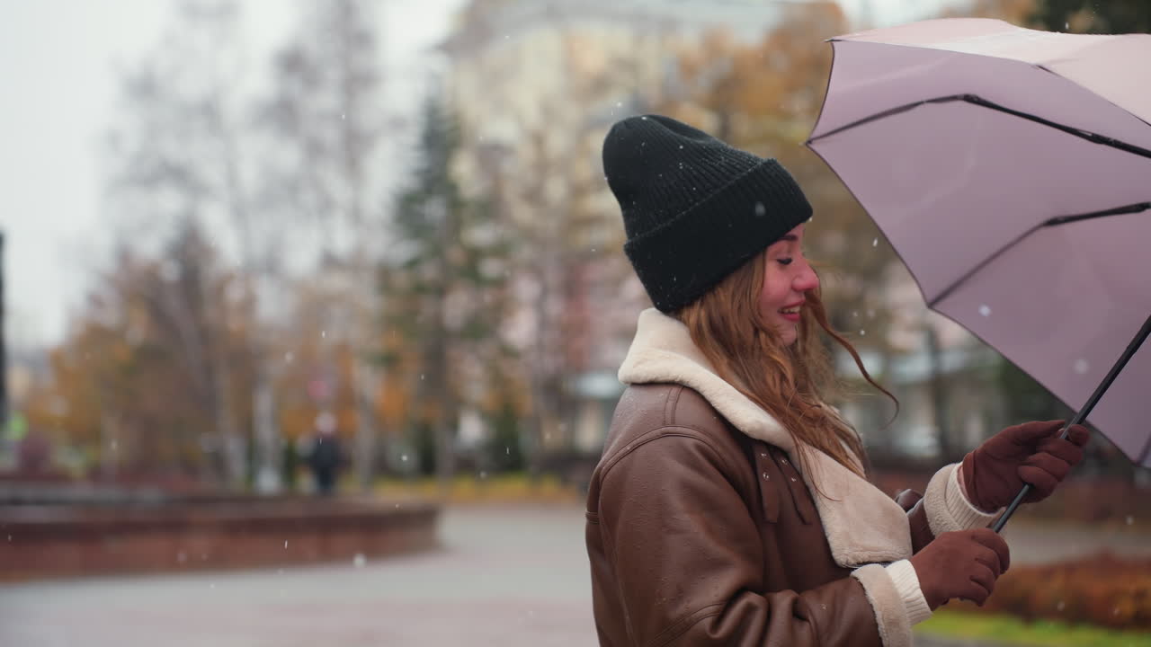 Cheerful lady holding umbrella, wearing knit cap, brown shearling jacket, happily spinning umbrella in light snowfall, surrounded by trees, enjoying autumn day, joyful and carefree moment outdoors