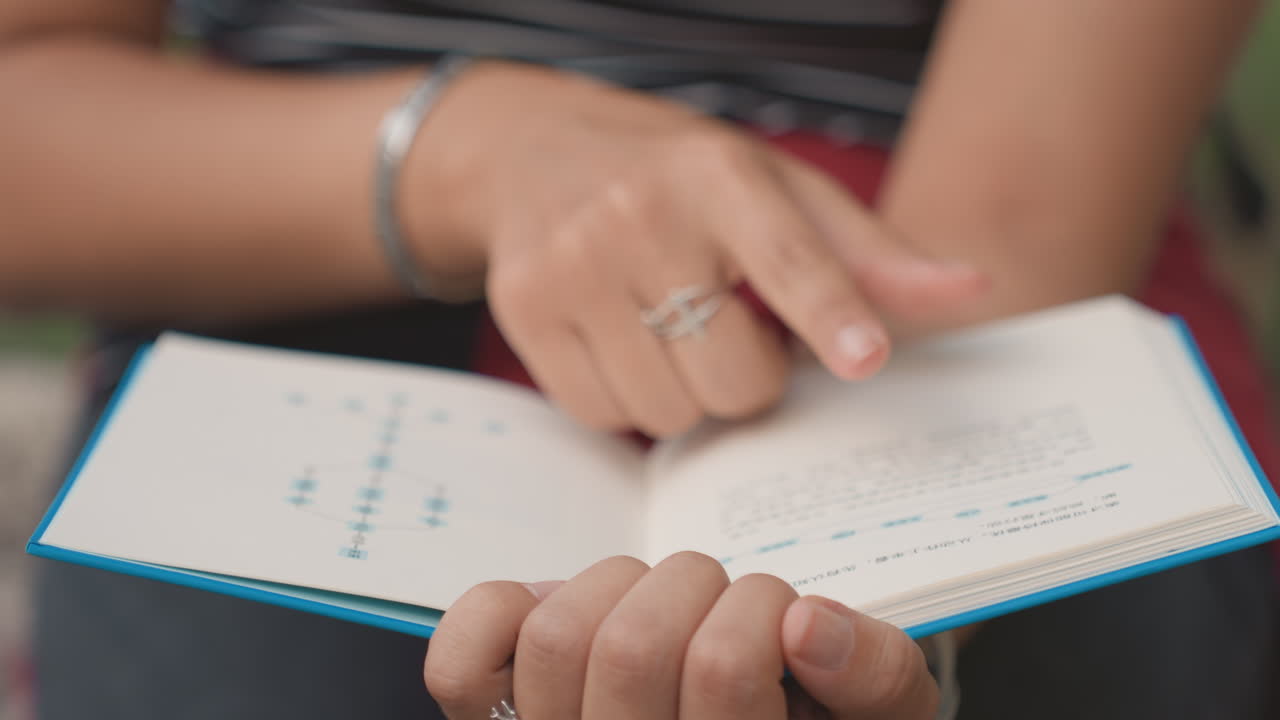 Closeup hands turning small book, ring and bracelet visible, finger tracing text on teal hardcover, outdoor bench setting with soft bokeh and sunlight, slow intimate reading ritual and focused study