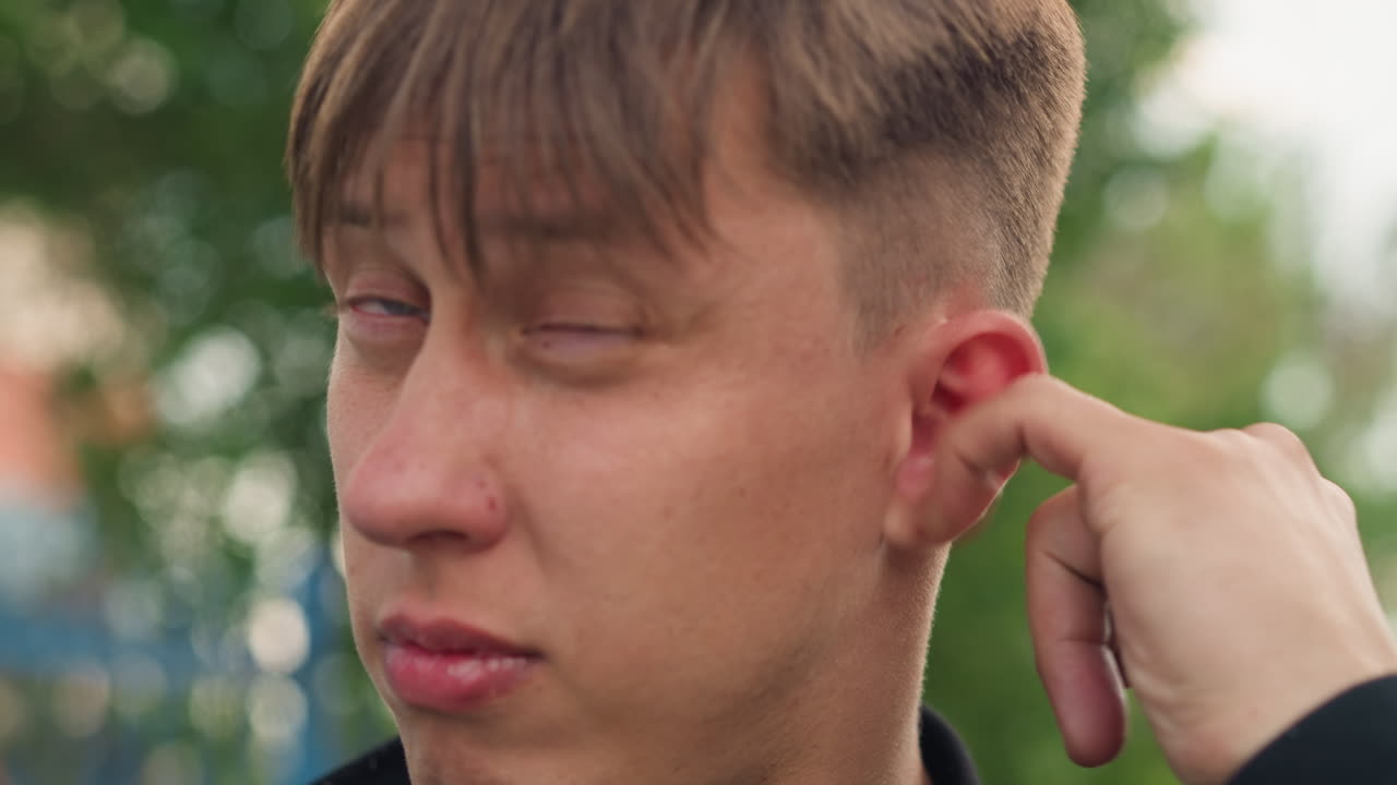 Молодой белый музыкант на улице проверяет наушник, жест проверки звука перед уличным выступлением, поправляет наушник-вкладыш и ушной монитор, взгляд сосредоточен вверх, лиственный фон и повседневная куртка