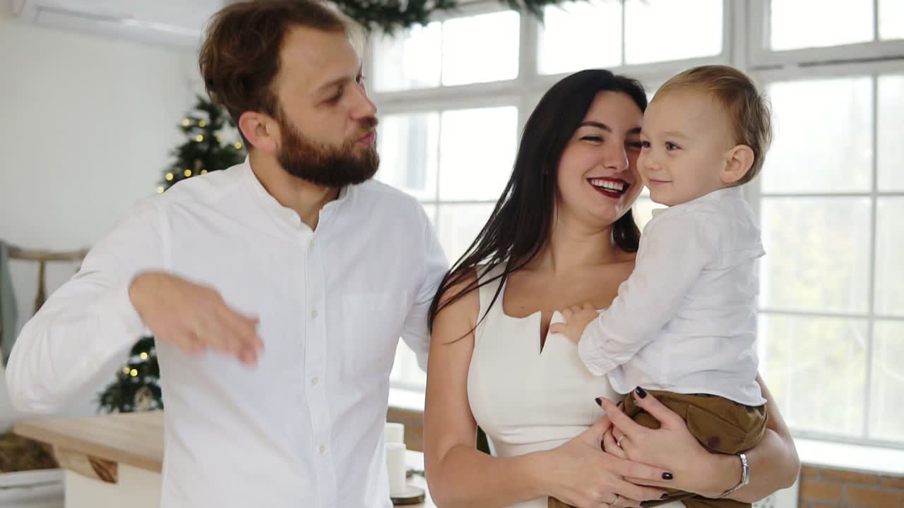 행복한 젊은 가족은 사랑스러운 아이를 손에 안고 크리스마스 트리 옆 세련된 아파트에서 함께 춤을 춥니다. 사랑스러운