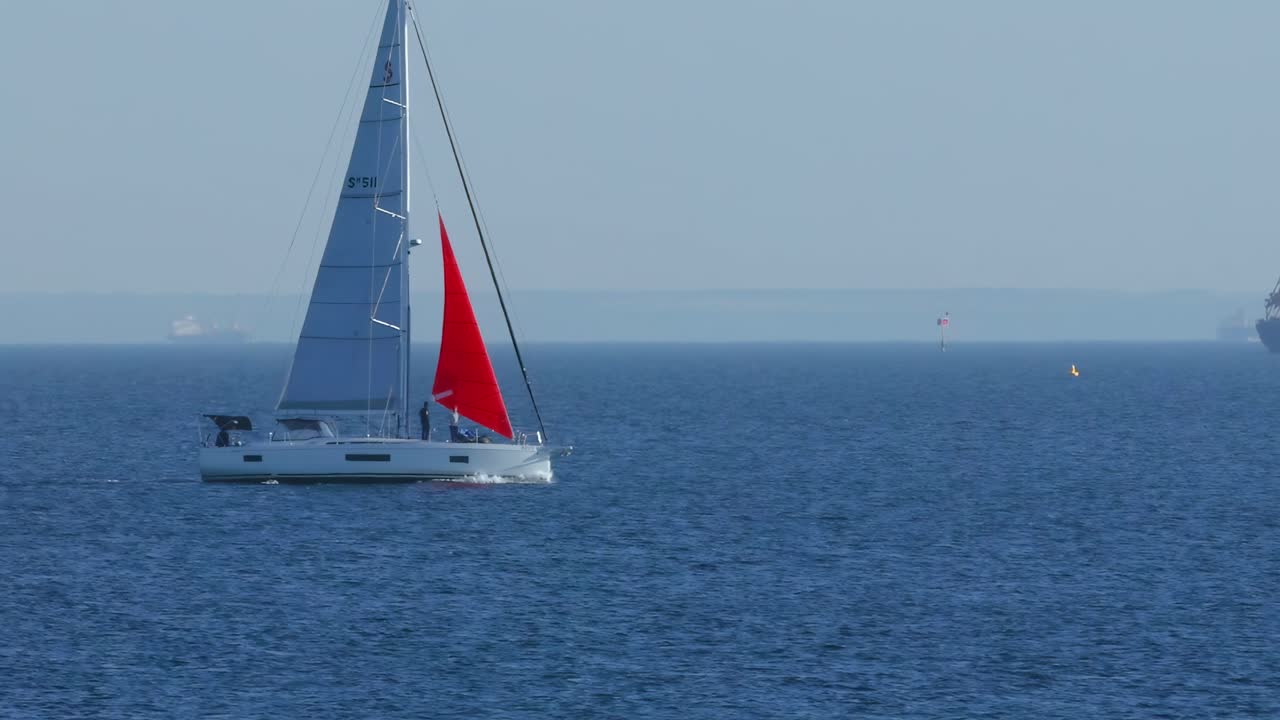 A sailboat with a striking red sail glides across the calm blue sea under a clear sky.