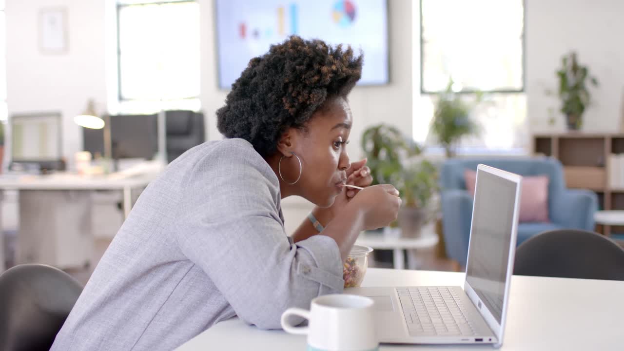 African american casual businesswoman eating lunch and using laptop in office, slow motion