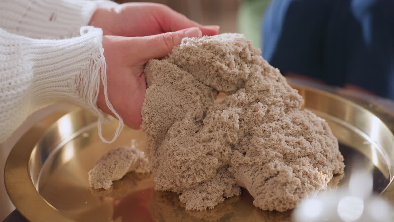 Close up view of female hands gently rubbing soft molded soil on golden tray during emotional healing session, capturing sensory therapy moment in peaceful indoor setting with warm lighting