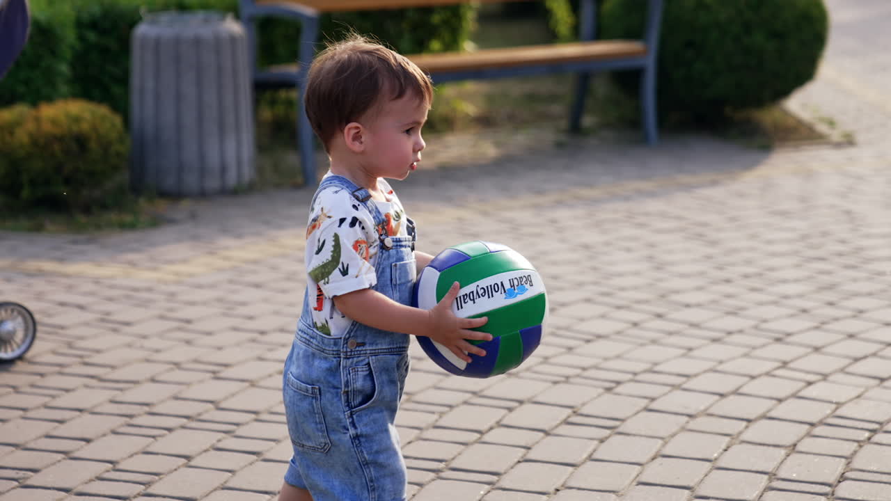 Adorable kid runs with a ball by the park in summer. Baby boy plays outdoors.