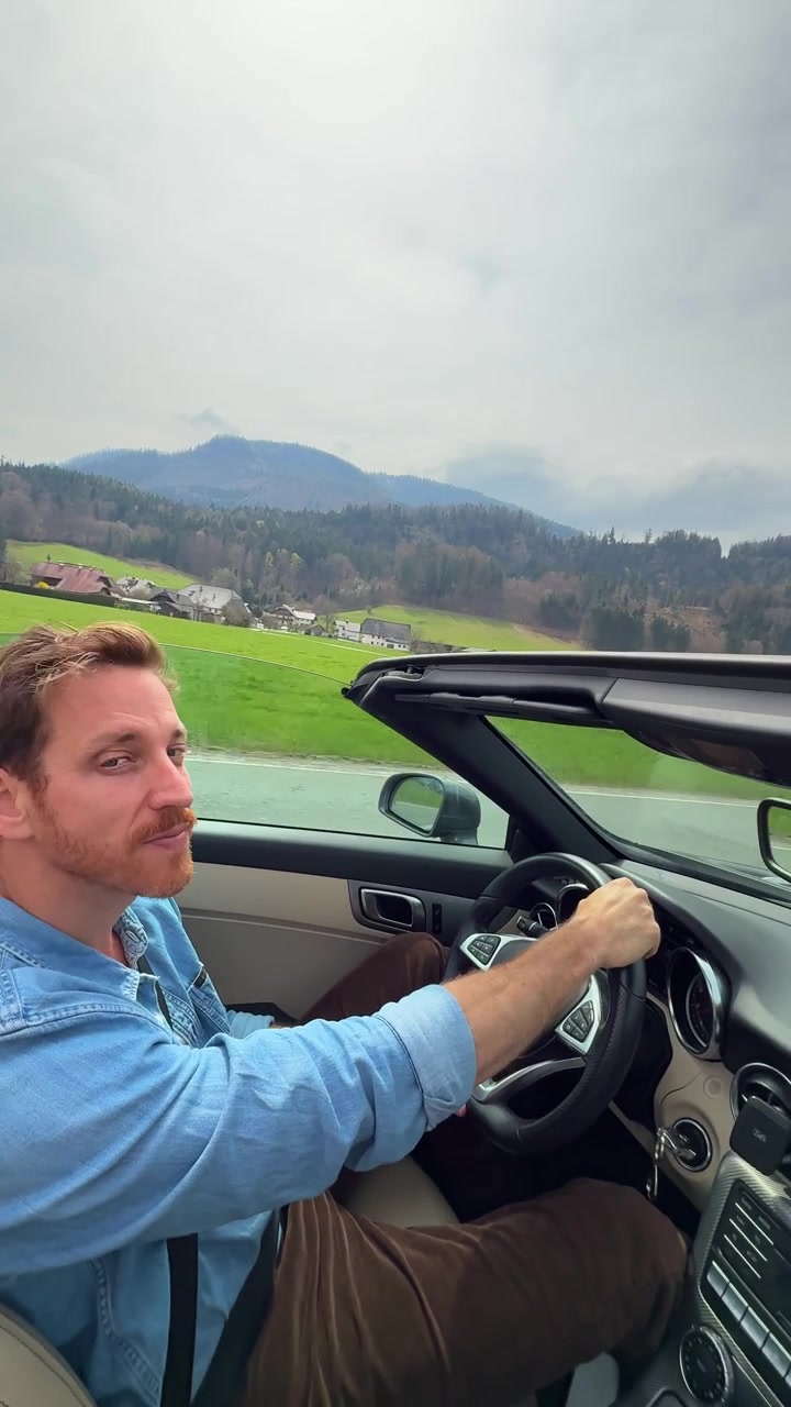 Young man enjoying a convertible drive through beautiful countryside roads with green fields and mountain scenery. Travel, freedom, road trip and adventure concept. Vertical lifestyle footage