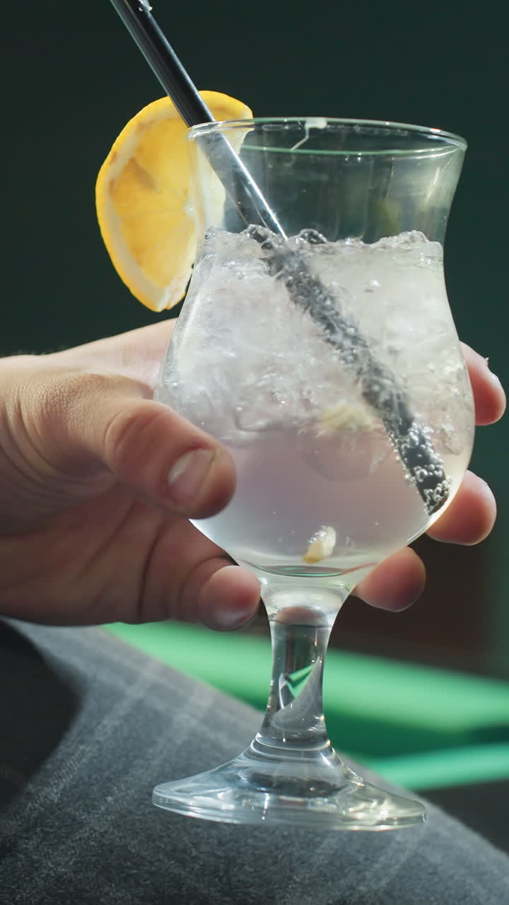 Primo piano di una persona, viso non visibile, che indossa una camicia bianca e pantaloni grigi, tiene in mano una stecca da biliardo e un bicchiere di limonata con cannuccia nera. Cubetti di ghiaccio e fetta di limone esaltano l'atmosfera rinfrescante.
