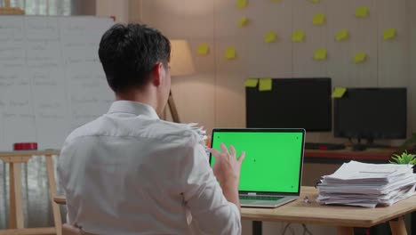vista trasera de un hombre asiático hablando con una computadora portátil de pantalla verde mientras trabaja con documentos en la oficina