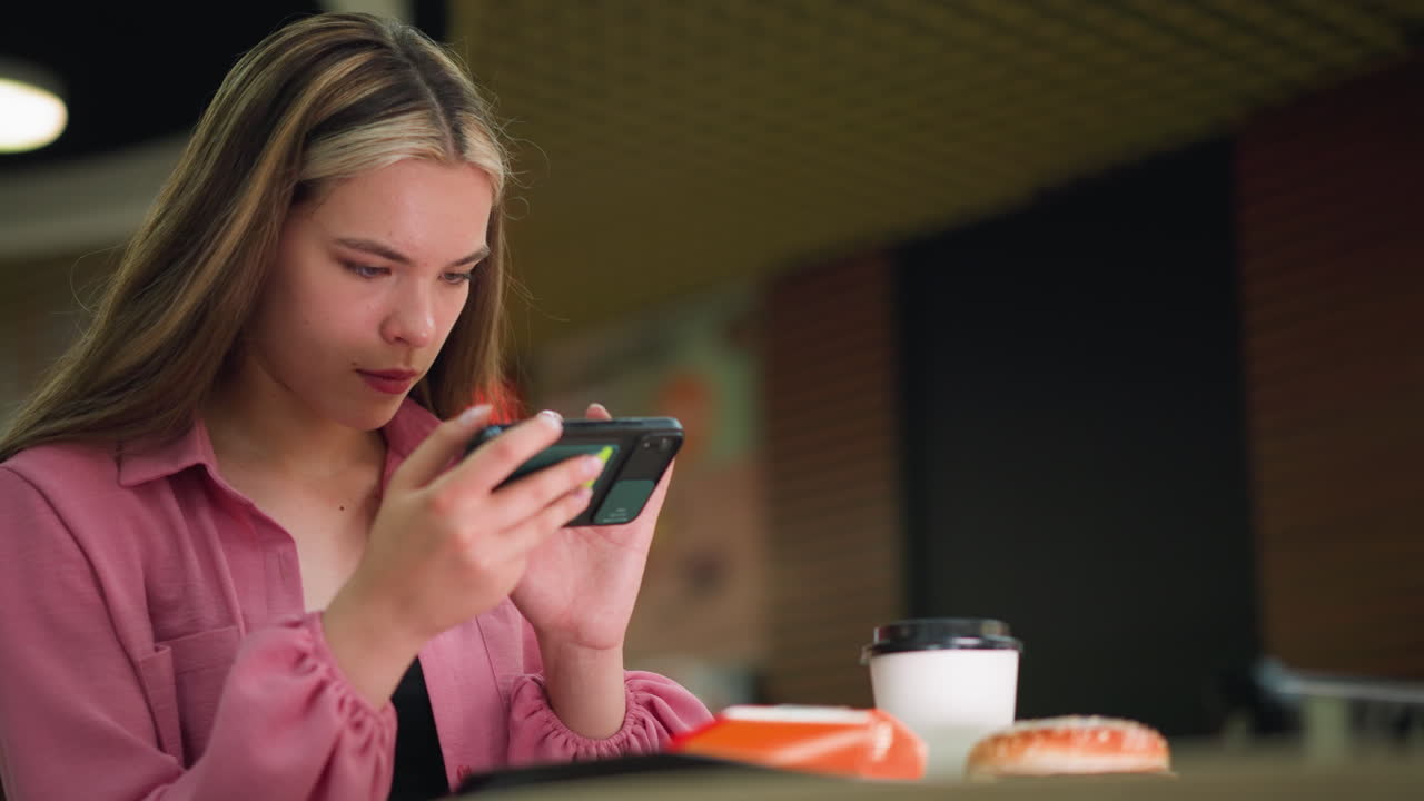 mujer en un vestido rosa centrándose atentamente en su teléfono, posiblemente tomando una foto de algo delante de ella, taza de café y hamburguesa en primer plano en la mesa, con un fondo borroso