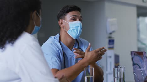 Mixed-race-male-doctor-and-female-hospital-colleague-wearing-face-masks-talking-at-meeting