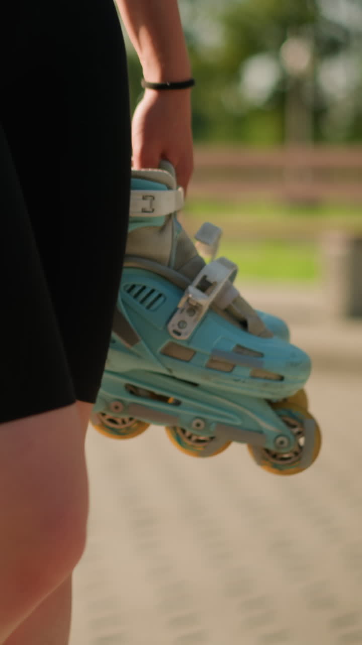 Lady wearing black tights walking outdoors while holding cyan roller skates in her right hand, with blurred background featuring benches, lush greenery, and a flower pot