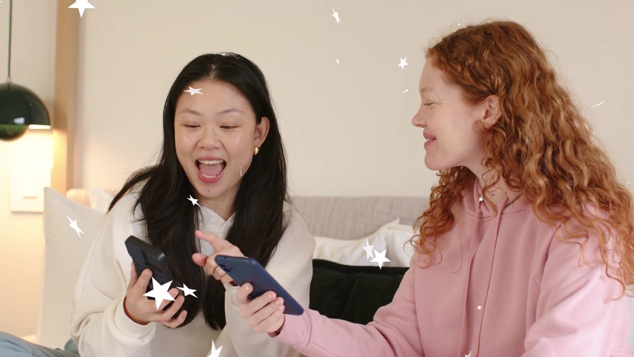 ベッドでテクノロジーを共有する2人の女性。右側が青いスマートフォンを差し出し、左側が暗いスマートフォンをタップしている。星のオーバーレイがかかっている。
