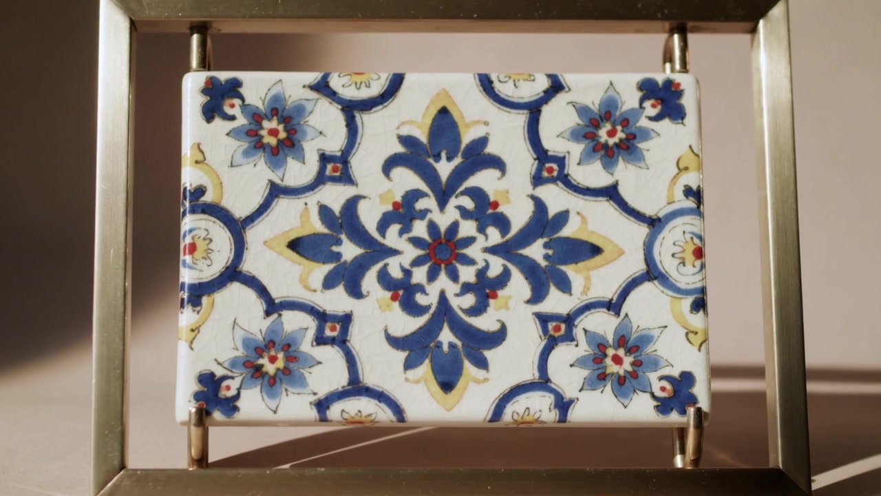 Telecamera che zooma e inizia la riproduzione, rivelando una piastrella in una cornice di metallo su un tavolo, con un motivo floreale blu.