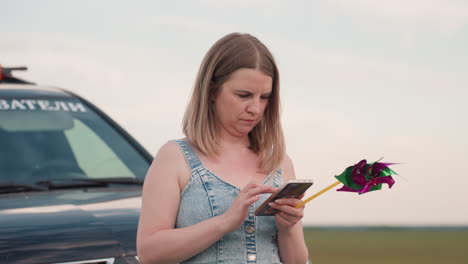 fair skin woman leans on black suv parked by country road using smartphone while holding vibrant pinwheel surrounded by open green fields and distant tree line under dramatic cloudy sunset sky