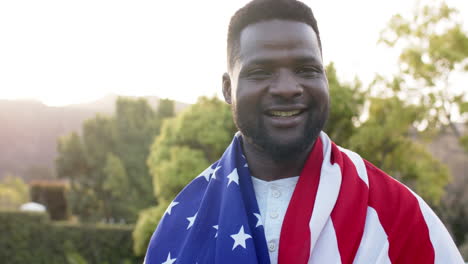 Happy-african-american-man-standing-with-flag-on-back-and-smiling-in-sunny-garden