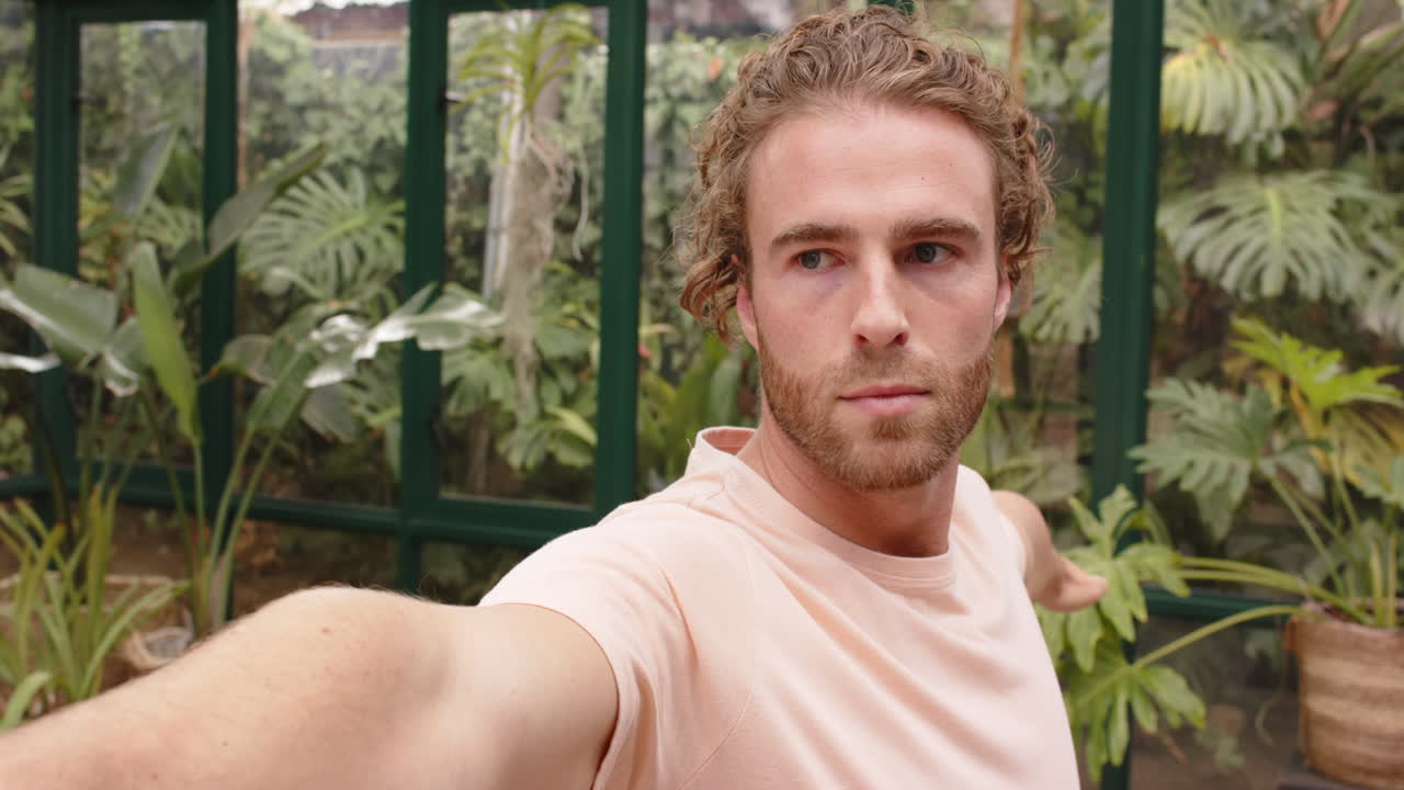 Practicing yoga, man stretching arm in glass house surrounded by plants