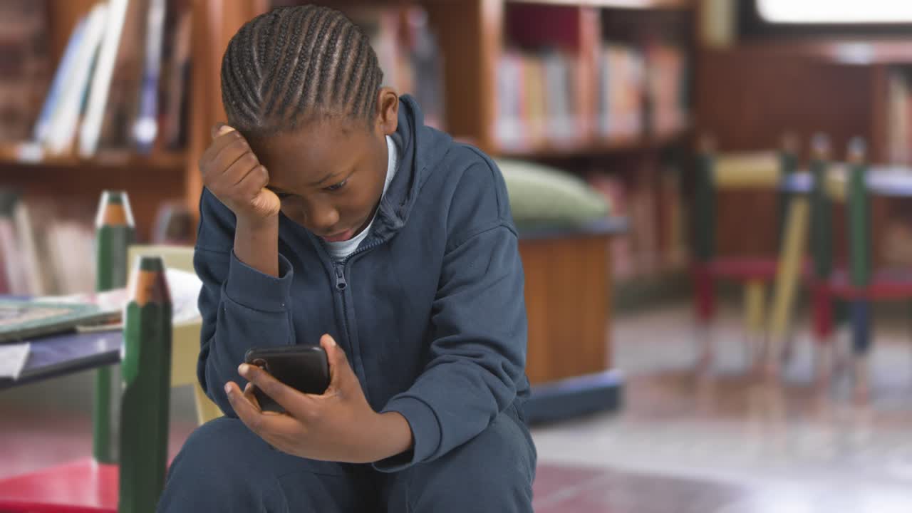 Unhappy Young Boy Being Cyberbullied On Mobile Phone In School Library